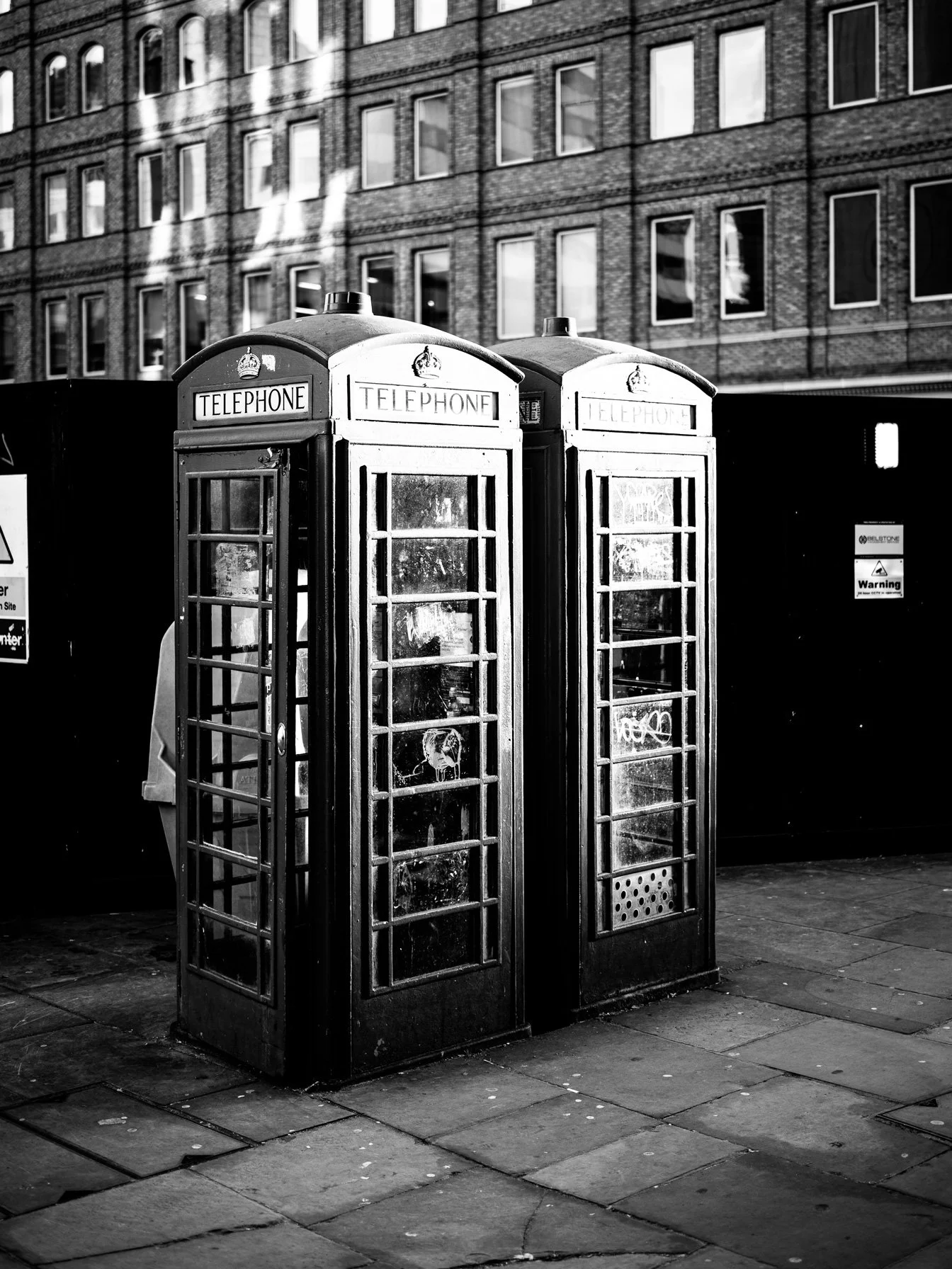 Two classic British telephone booths on a city sidewalk in black and white with a brick building in the background.