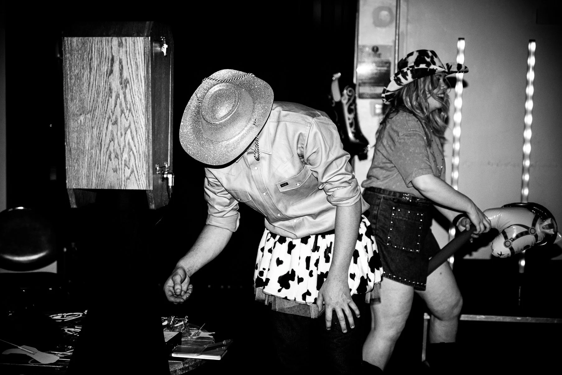 Two women in cowboy hats, one with a cow-print skirt, are at an arcade or amusement venue. One is holding onto a pogo stick and the other is leaning on the pogo stick.