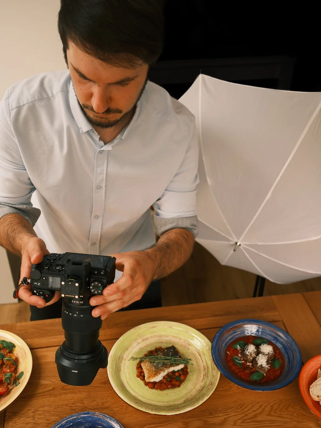A man in a white shirt is taking a photo of plated dishes on a wooden table, with a white umbrella lighting setup behind him.