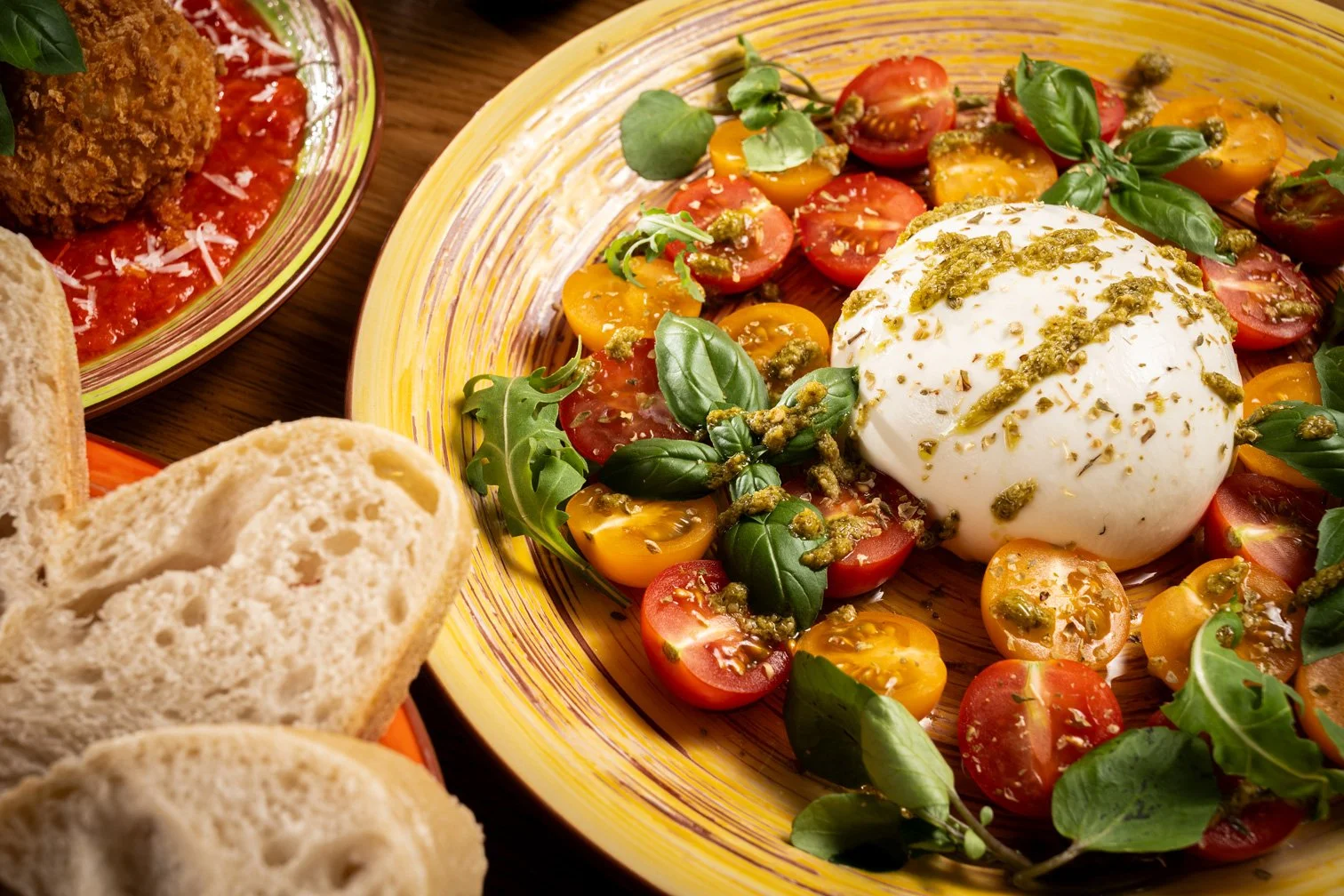 Caprese salad with cherry tomatoes, fresh basil, mozzarella, and pesto on a yellow plate, with slices of bread and a dish of breaded fried food with tomato sauce on a wooden table.