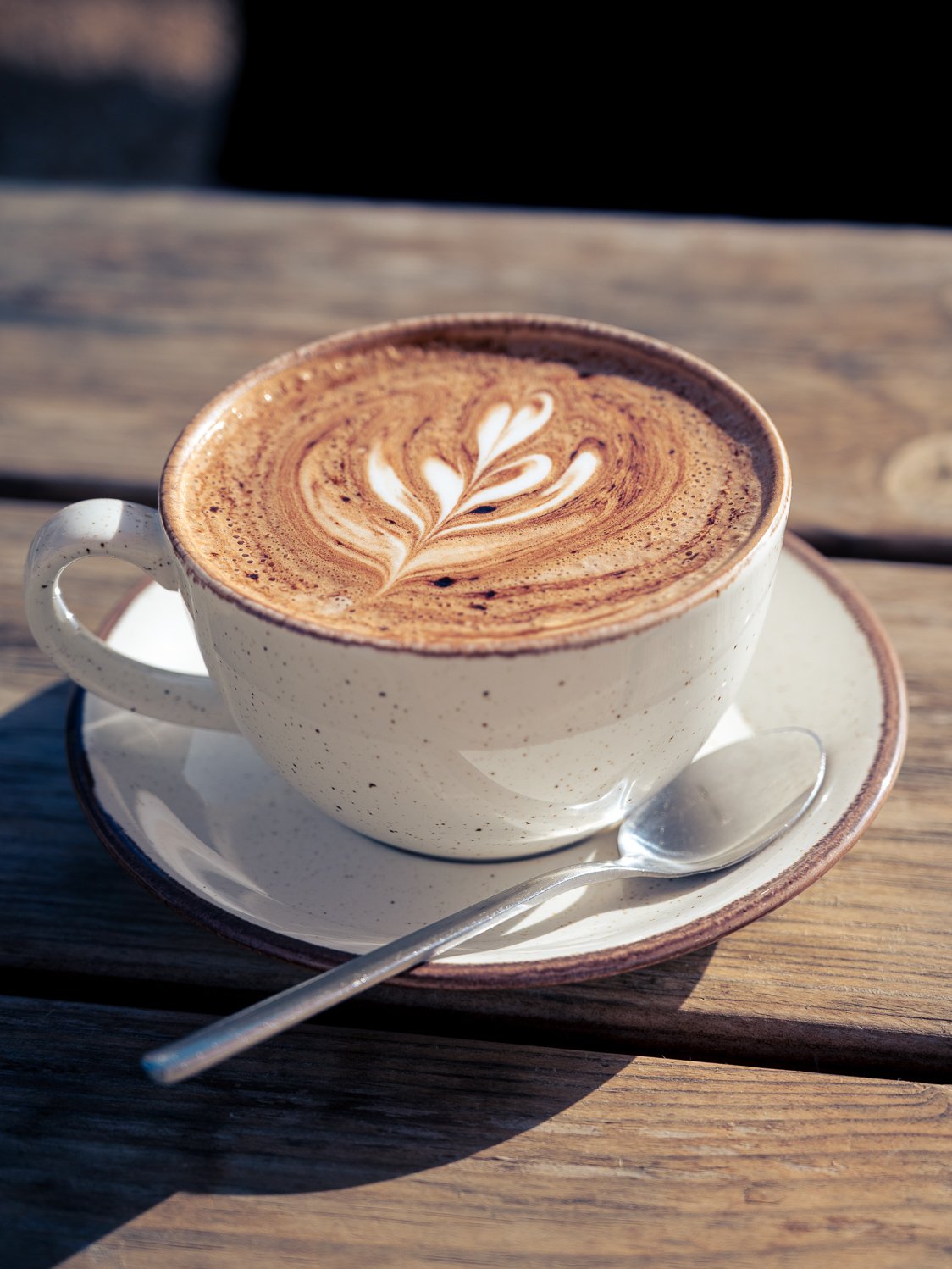 A cup of coffee with latte art on top, on a saucer with a spoon, placed on a wooden table.