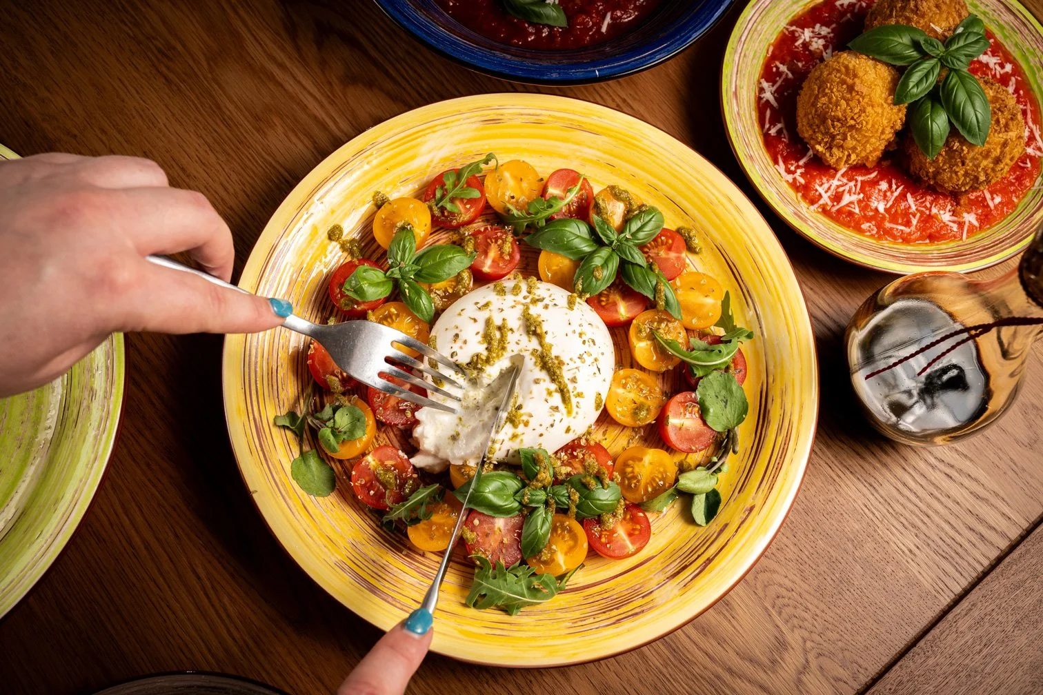 A salad with cherry tomatoes, fresh basil, and a large ball of mozzarella cheese on a yellow plate, with a person cutting into the cheese with a fork and knife. Nearby are bowls of fried food in tomato sauce and a glass of dark soda.