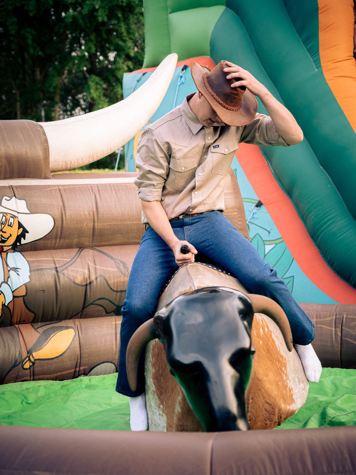 Person wearing cowboy hat sitting on a mechanical bull at a fair or carnival, with a colorful inflatable slide in the background.