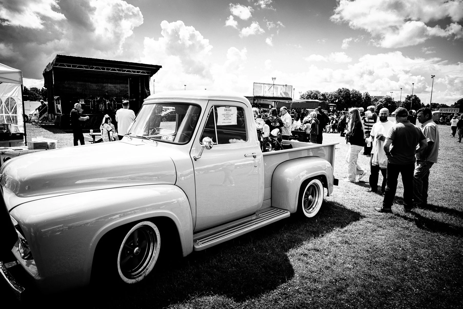 A classic vintage pickup truck parked on grass at a daytime outdoor event, with a crowd of people and a stage in the background.