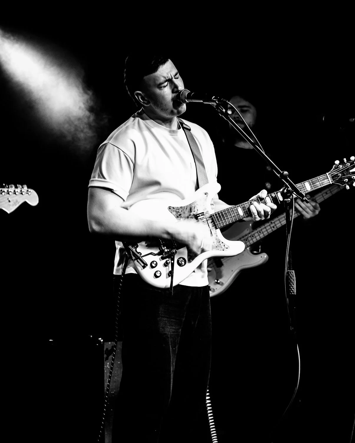A black and white photo of a man singing into a microphone while playing an electric guitar during a performance, with a bassist in the background.