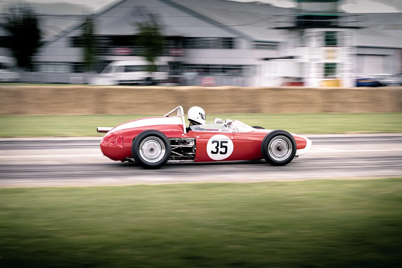 A vintage red race car with the number 35 on the side driving on a racetrack, with a blurry background of buildings and trees.