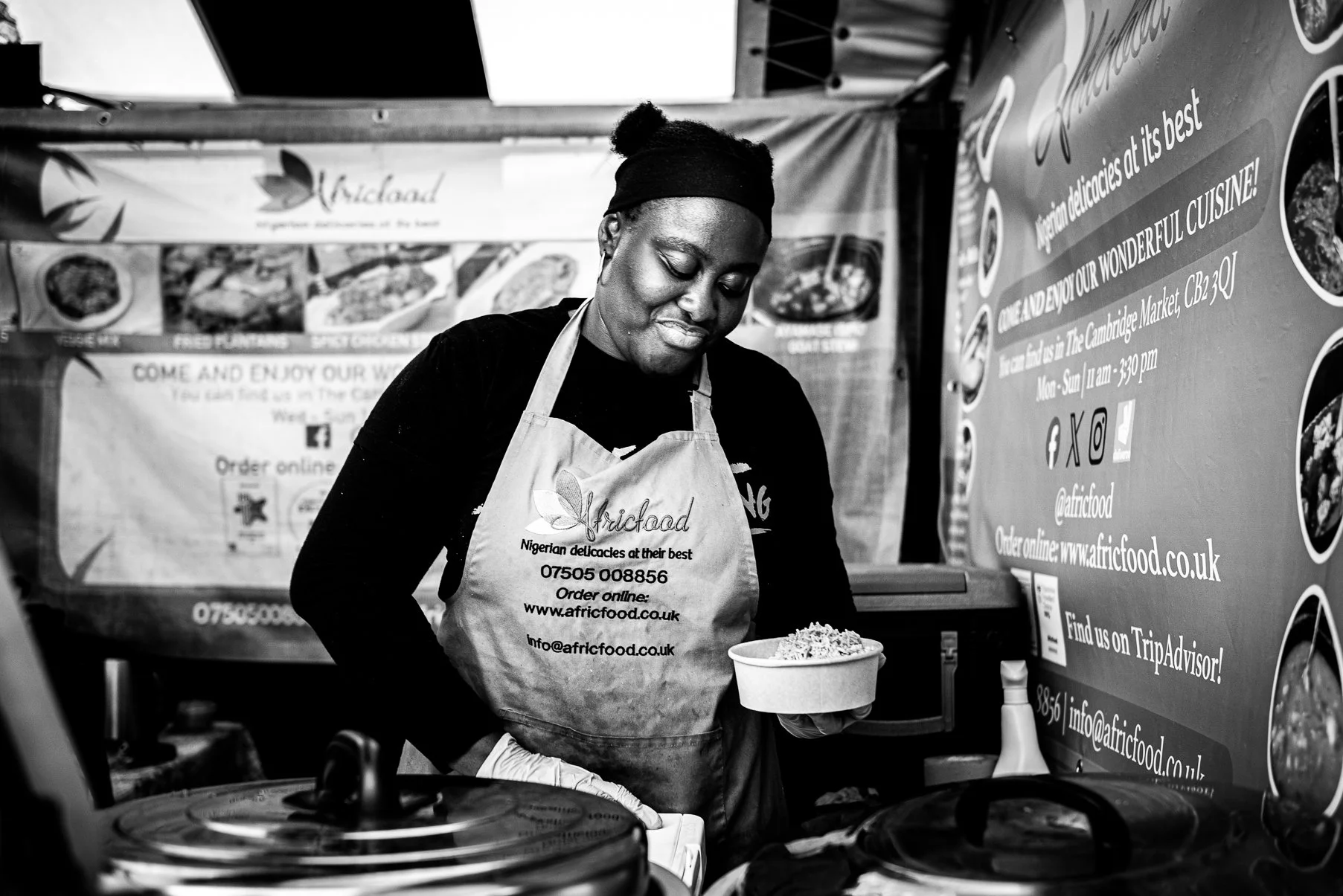 A woman working at a food booth, wearing an apron and holding a bowl of food, with promotional signs and menus displayed behind her.