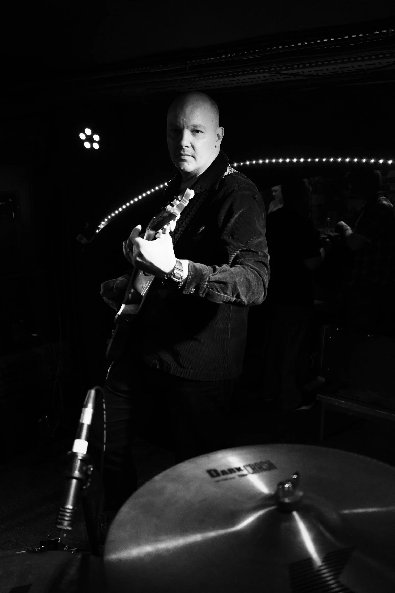 A black and white photo of a bald man holding a guitar, looking towards the camera with a serious expression, in a dimly lit room with stage lights and a drum set in the foreground.