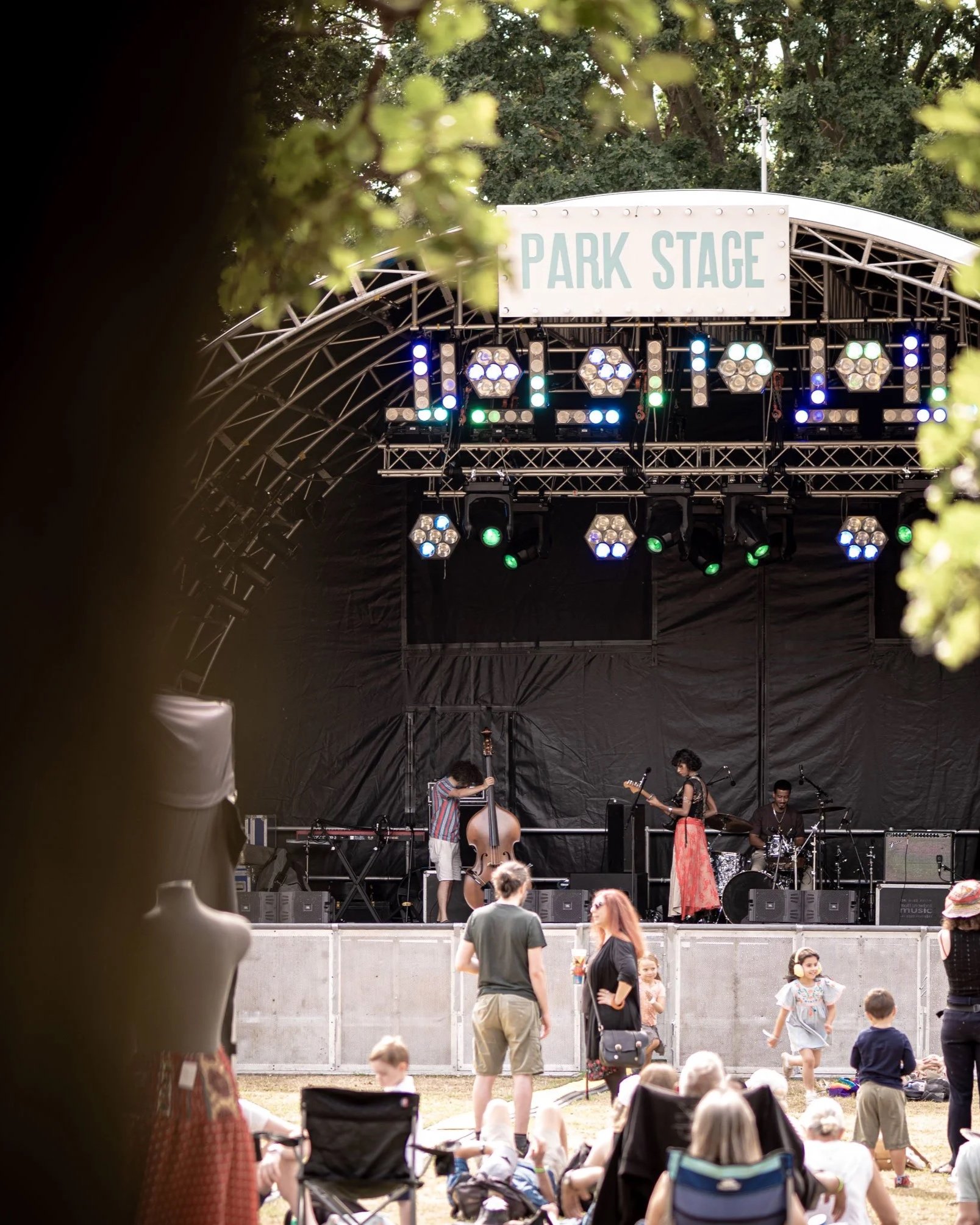 Children and adults watching a live musical performance on a stage at an outdoor park festival, with the stage labeled 'PARK STAGE' and various stage lights.