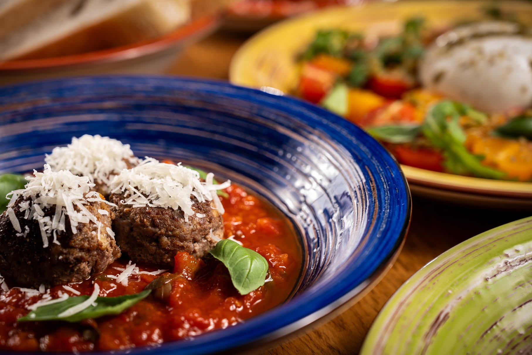 Close-up of a traditional Italian dish with meatballs topped with grated cheese in a red tomato sauce, garnished with basil leaves, served in a blue hand made ceramic bowl.