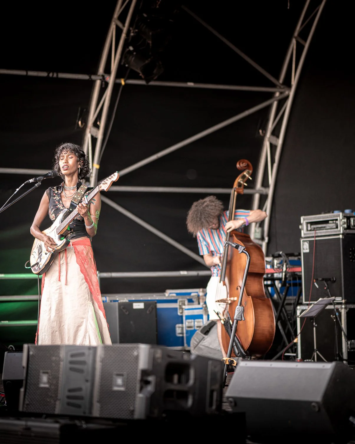 Two musicians performing on an outdoor stage: a woman singing and playing electric guitar, and a man playing upright bass, with stage equipment and scaffolding in the background.
