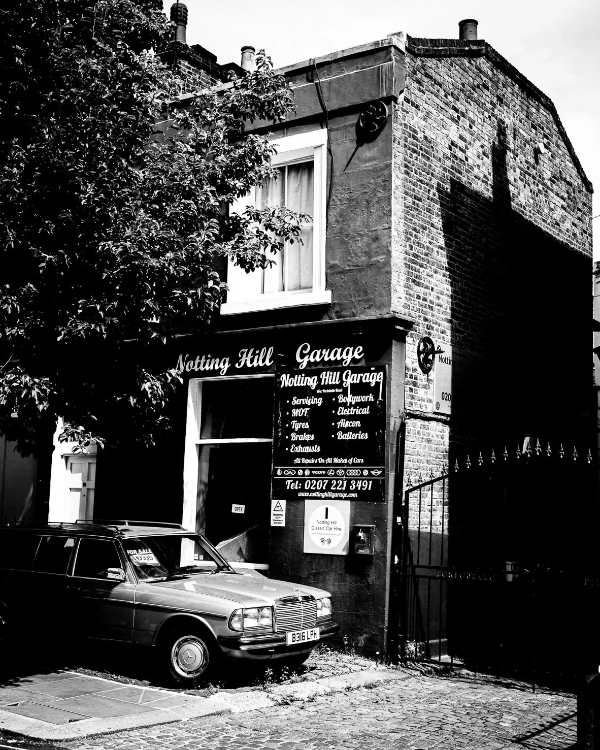 Black and white photo of a two-story brick building with a garage sign reading "Notting Hill Garage". A car is parked in front with a "For Sale" sign. The garage offers services like servicing, MOT, tyres, brakes, exhausts, electrical, aircon, and ba