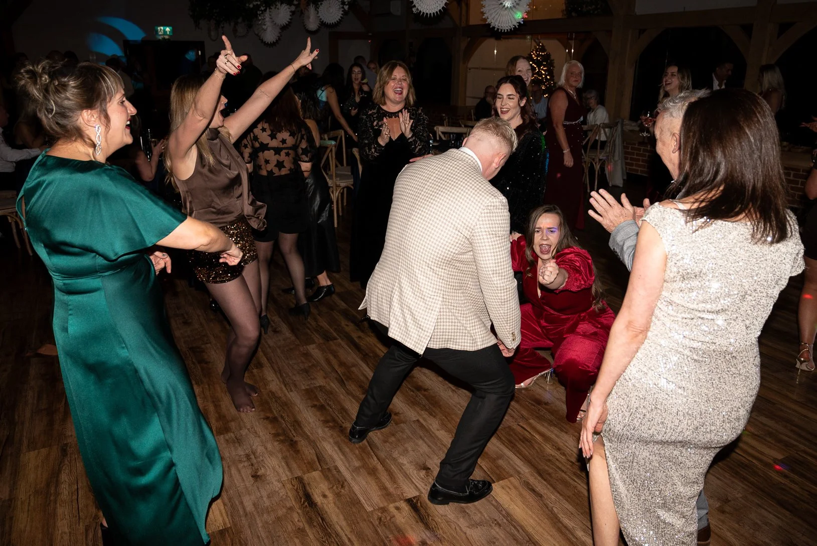 People dancing and celebrating at a party, with some laughing and cheering, in a decorated venue with paper lanterns and a Christmas tree in the background.