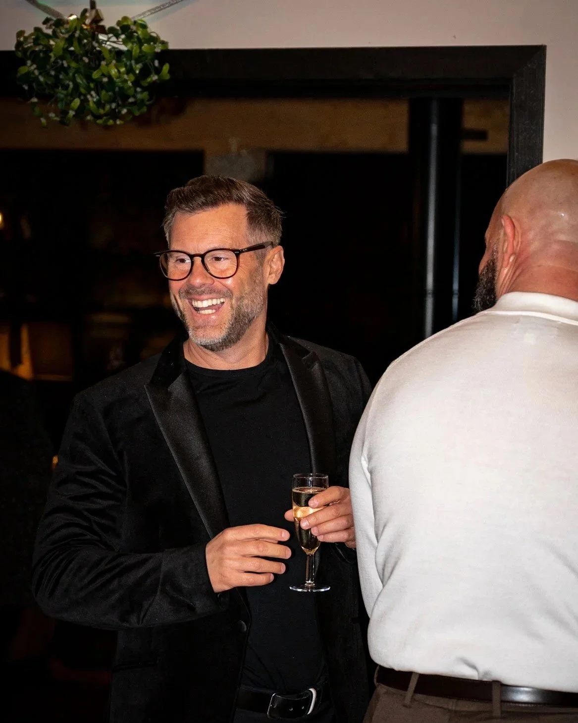 A man with glasses and a beard smiling and holding a glass of champagne, engaging in conversation with another man at a corporate gathering.