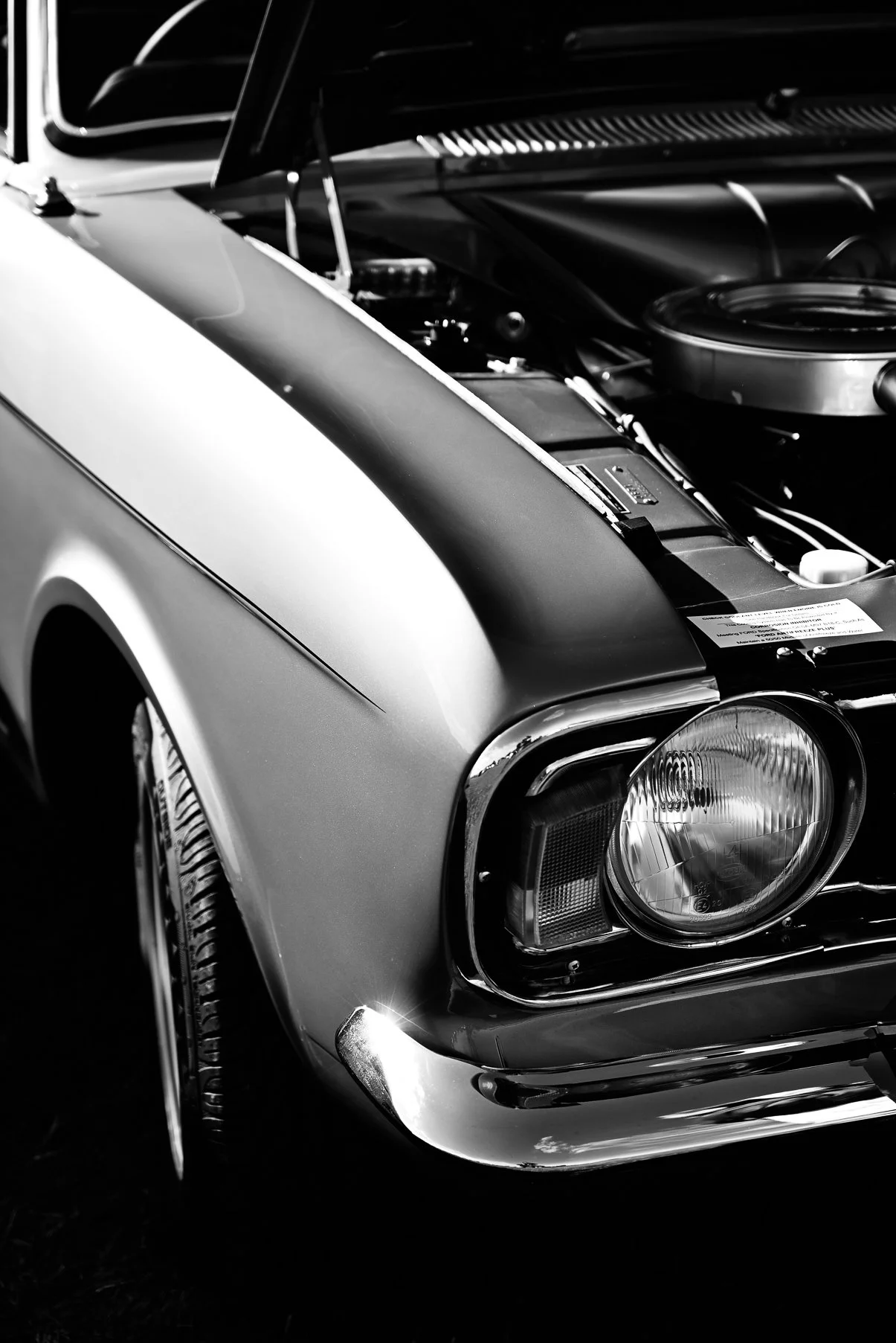 Black and white close-up of a vintage car's front end, showing the headlight, tire, and part of the hood with the hood open, revealing engine components.