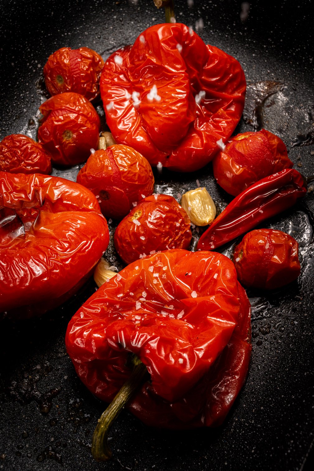 Charred and roasted red tomatoes, peppers and chilli peppers cooking in a black skillet with pieces of garlic.