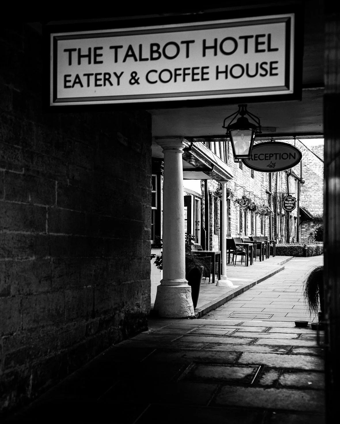 Black and white photo of the exterior of The Talbot Hotel, showing a pub sign, a porch with a lantern, outdoor seating, and a street view.