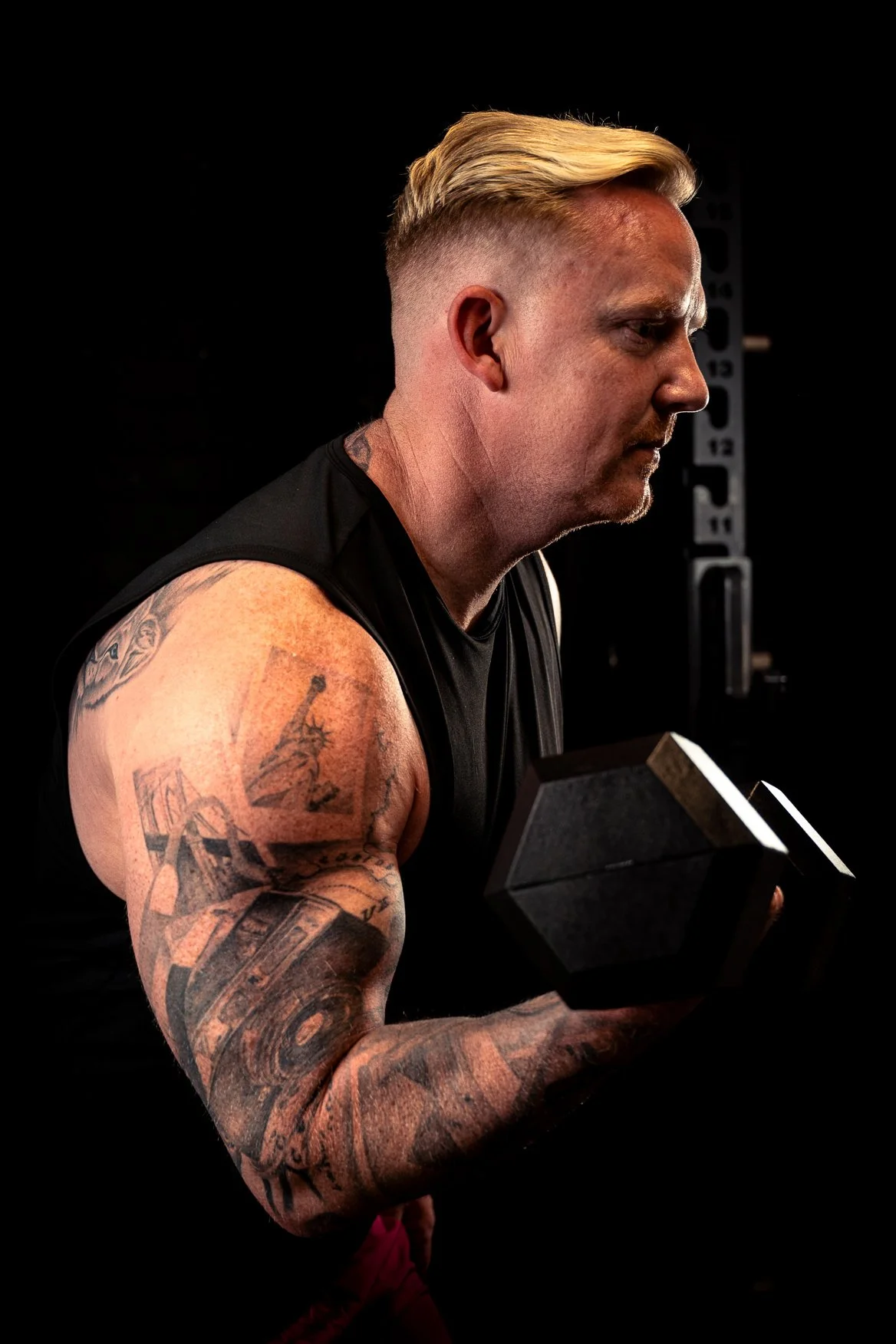 A muscular man with tattoos on his arm, wearing a sleeveless black shirt, is lifting a dumbbell in a dark gym setting.
