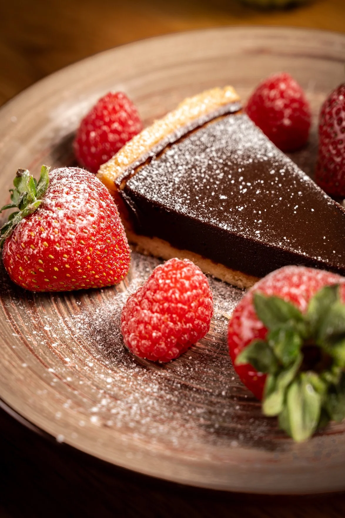 A slice of chocolate cake with powdered sugar on top, surrounded by strawberries and raspberries on a wooden plate.