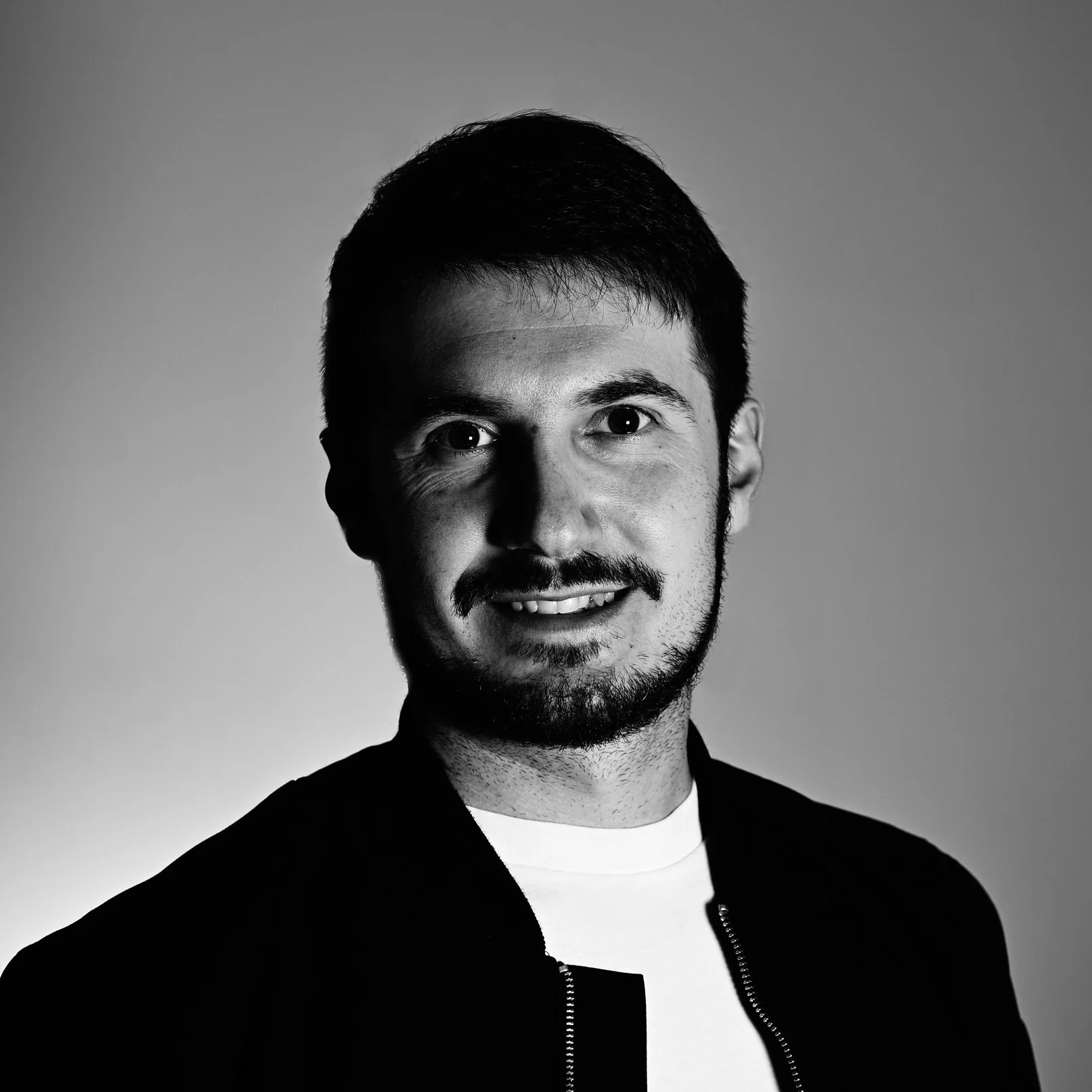 Black and white portrait of a man with short dark hair, facial hair, and a friendly expression, wearing a black jacket over a white shirt.