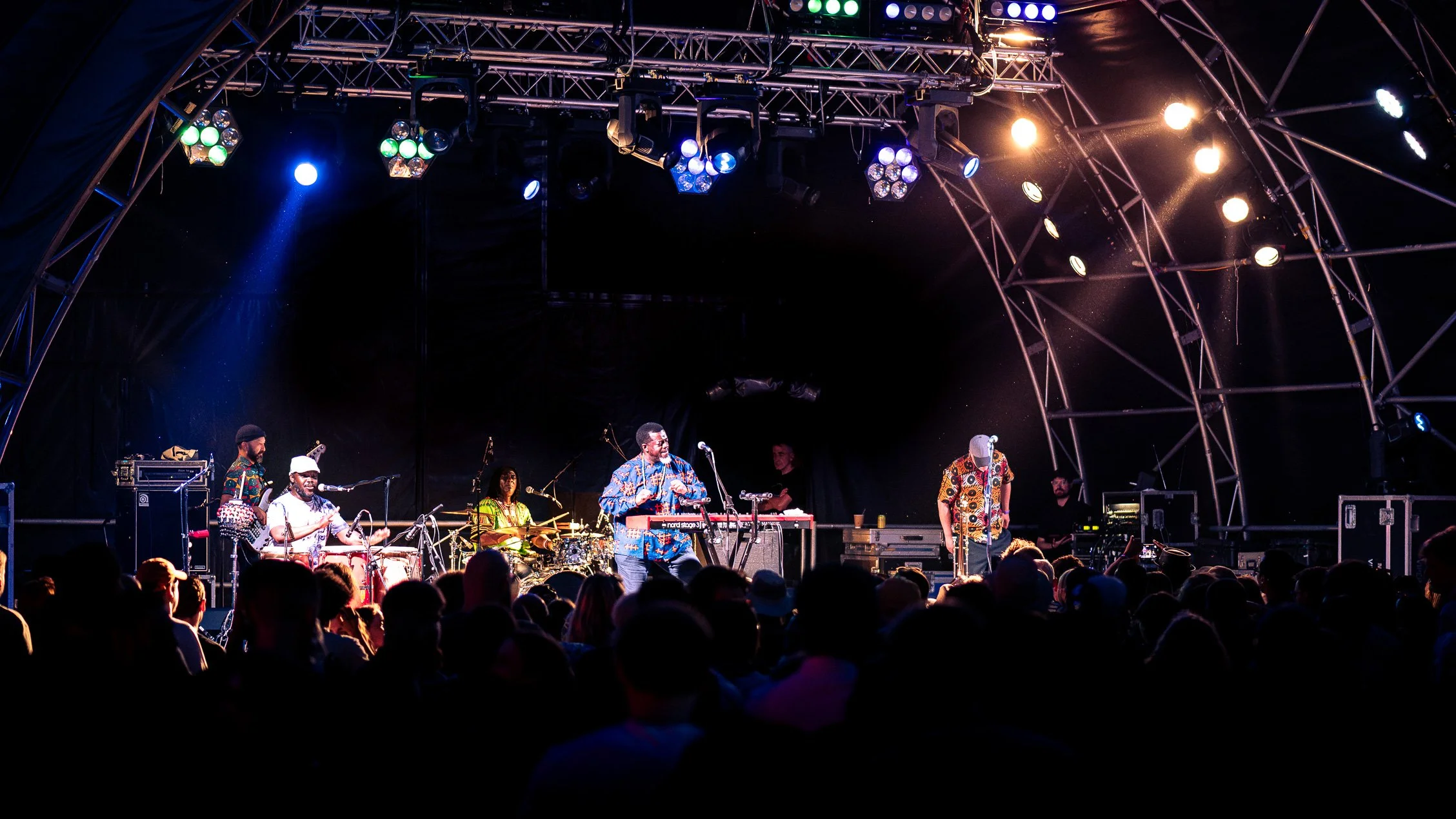 A live concert stage with musicians performing, surrounded by audience, under stage lighting.