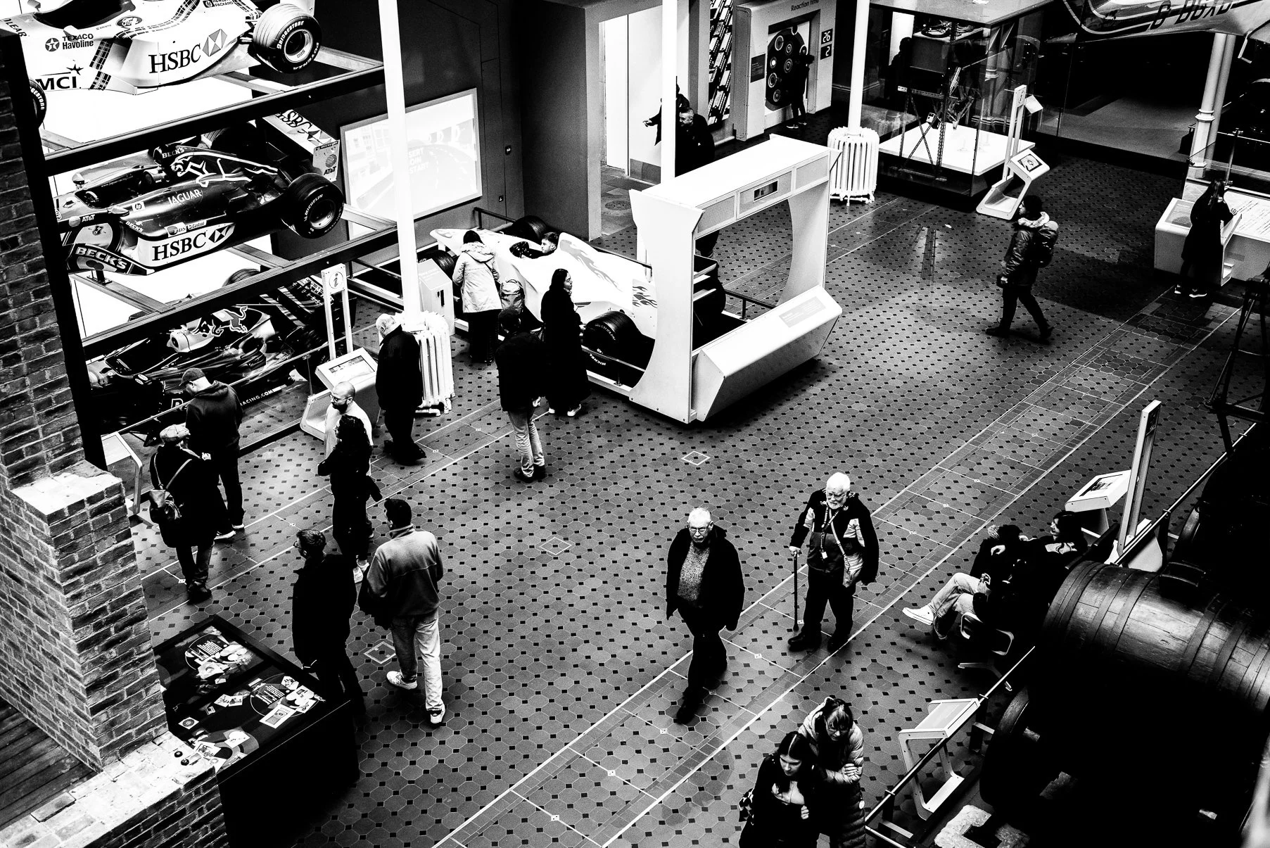 Black and white photo of an indoor exhibition at a museum with people browsing display cases and racing car models on glass shelves, with some seated and others walking around.