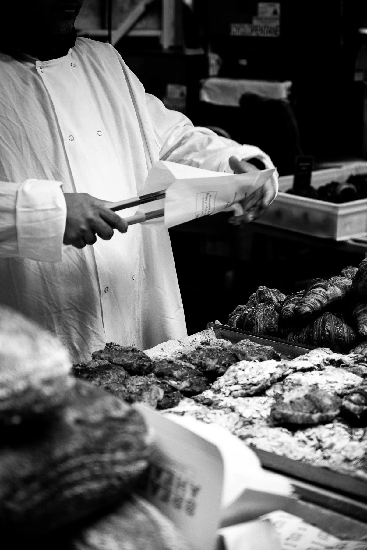 A chef in a white coat is handling a piece of paper or a menu, standing near a display of baked goods, possibly in a bakery or a kitchen.