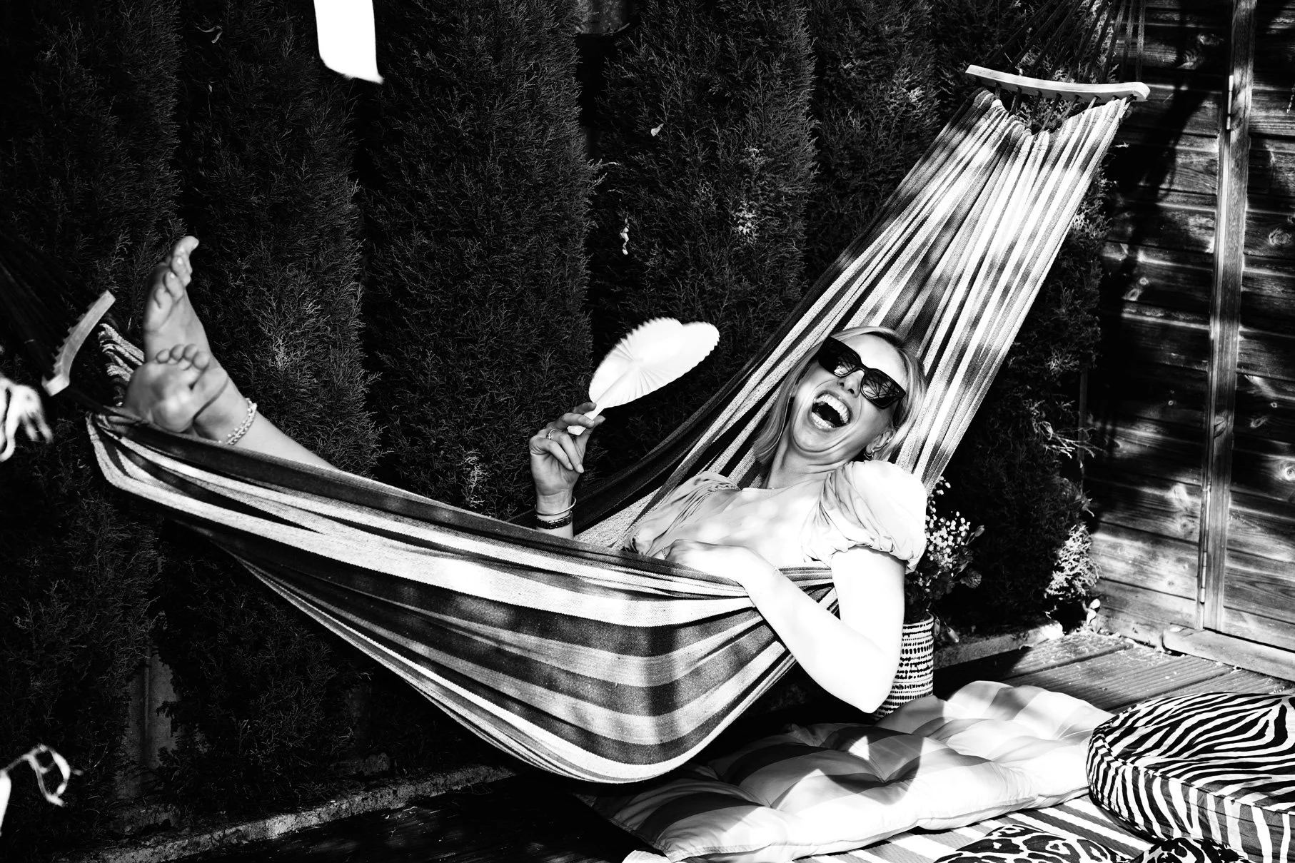 A woman smiling and laughing while lying in a hammock outdoors, wearing sunglasses and holding a small umbrella, with bushes and a wooden fence in the background.
