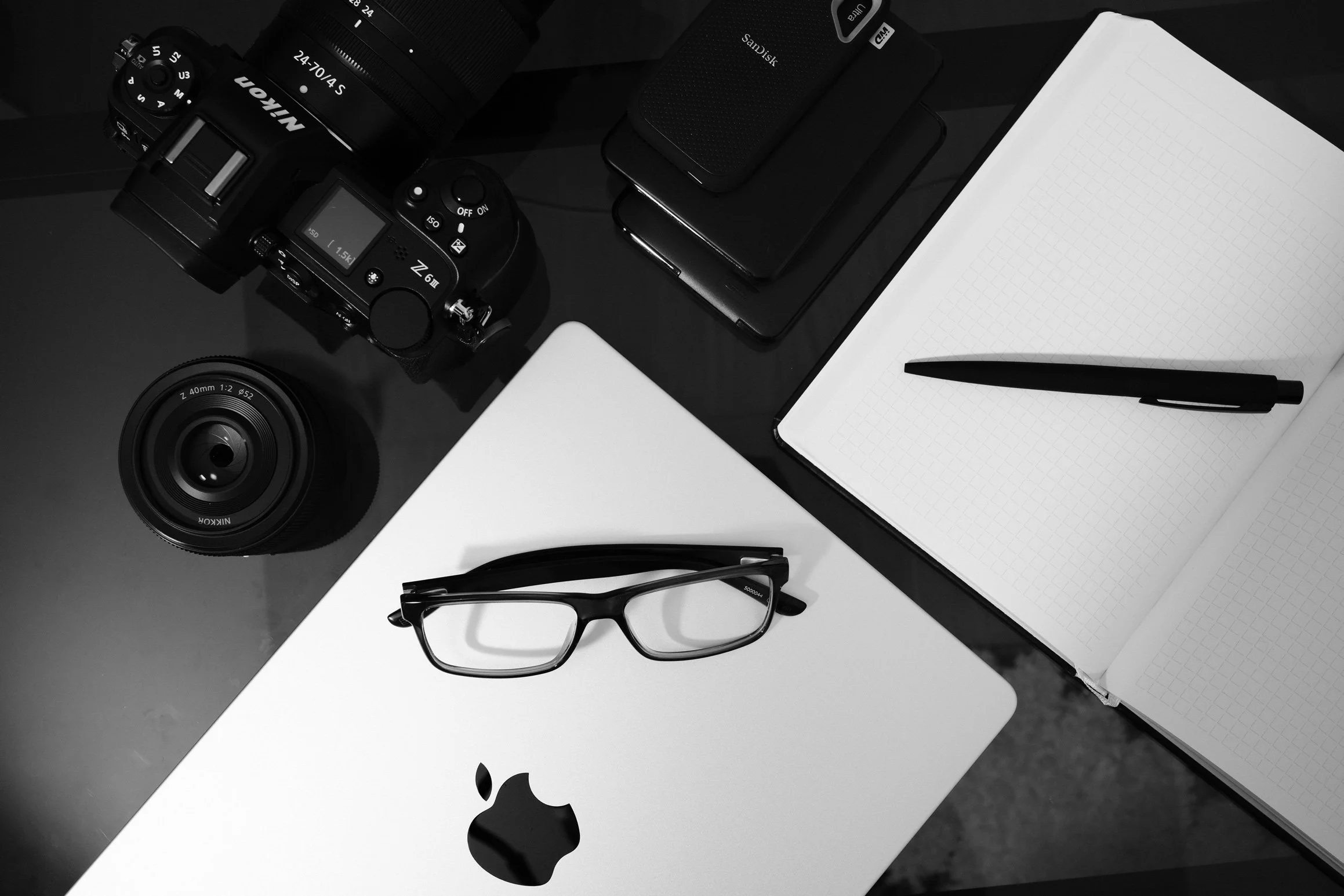 Black and white photograph of a workspace with a camera, glasses, a closed laptop, notebooks, a pen, a smartphone, and a desktop computer.