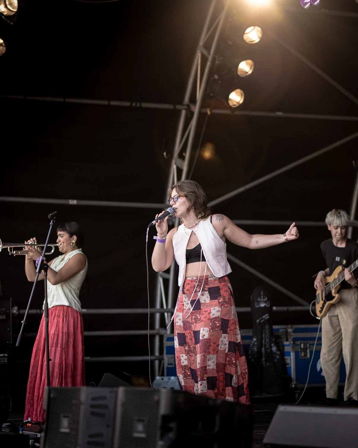 Three musicians performing on a stage. The woman in the center sings into a microphone, has short brown hair, glasses, a sleeveless vest, a black crop top, and a patchwork long skirt. To her left, a woman plays the trumpet, wearing a sleeveless top a