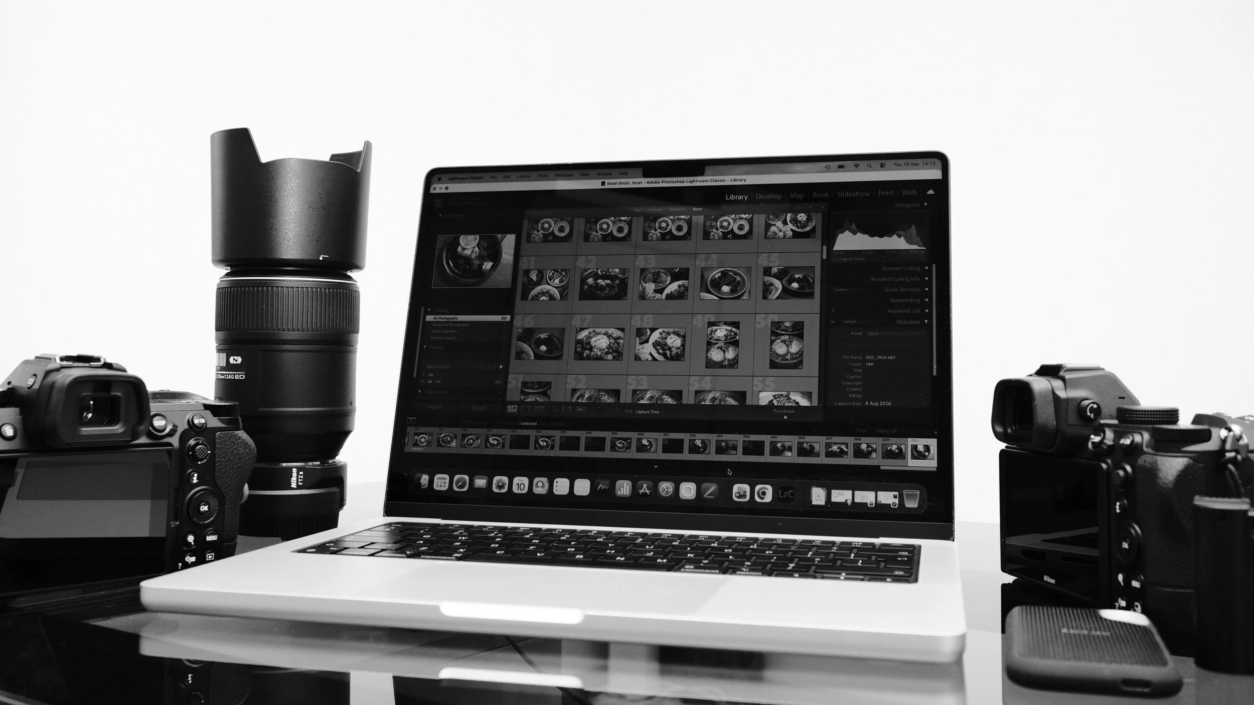 A workspace with a camera, a lens, a laptop displaying photo editing software, and several camera bodies on a desk.