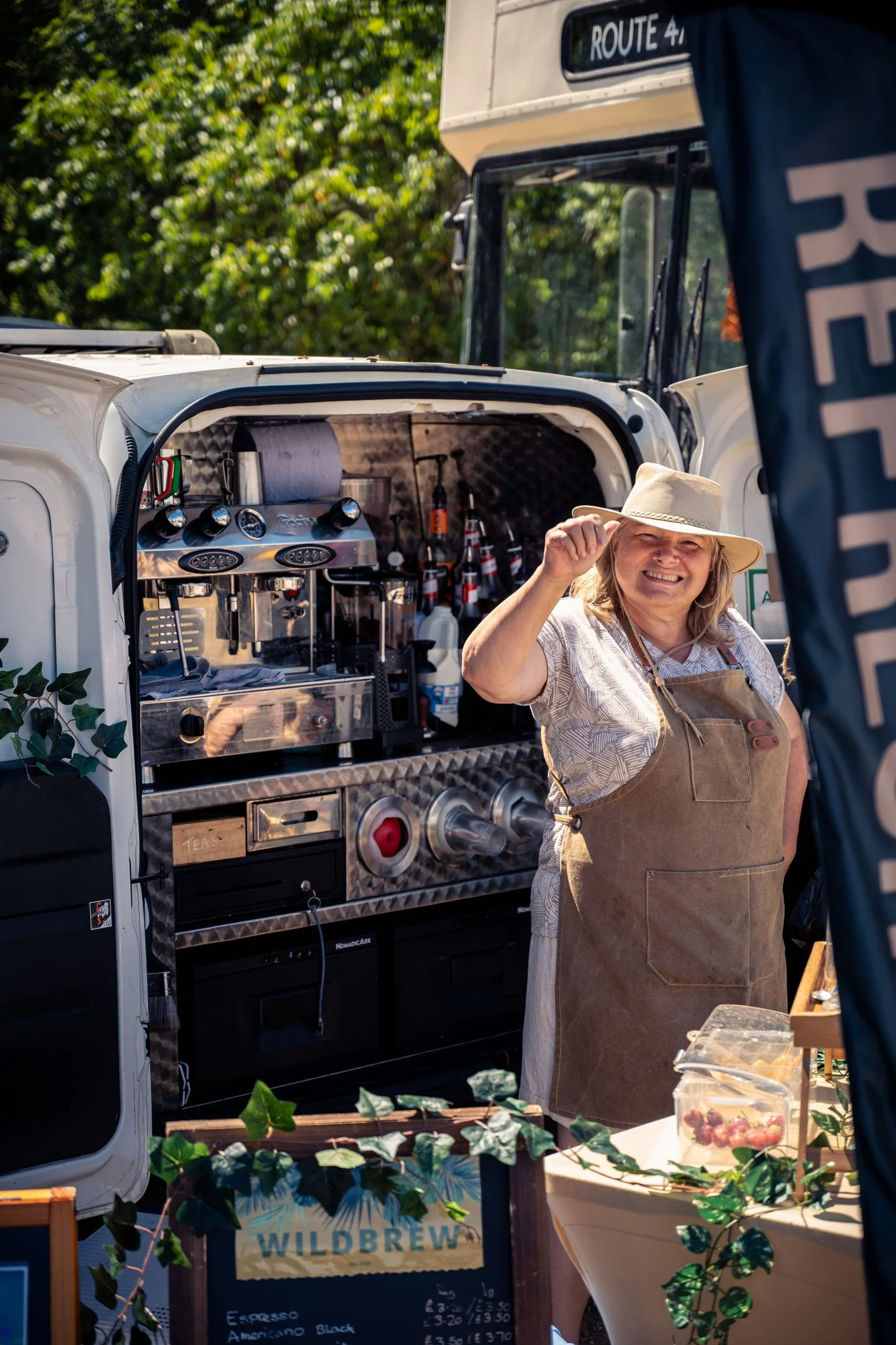 A smiling woman wearing a wide-brimmed hat and apron standing next to a coffee cart with a menu for espresso and American Black coffee, with greenery in the background.