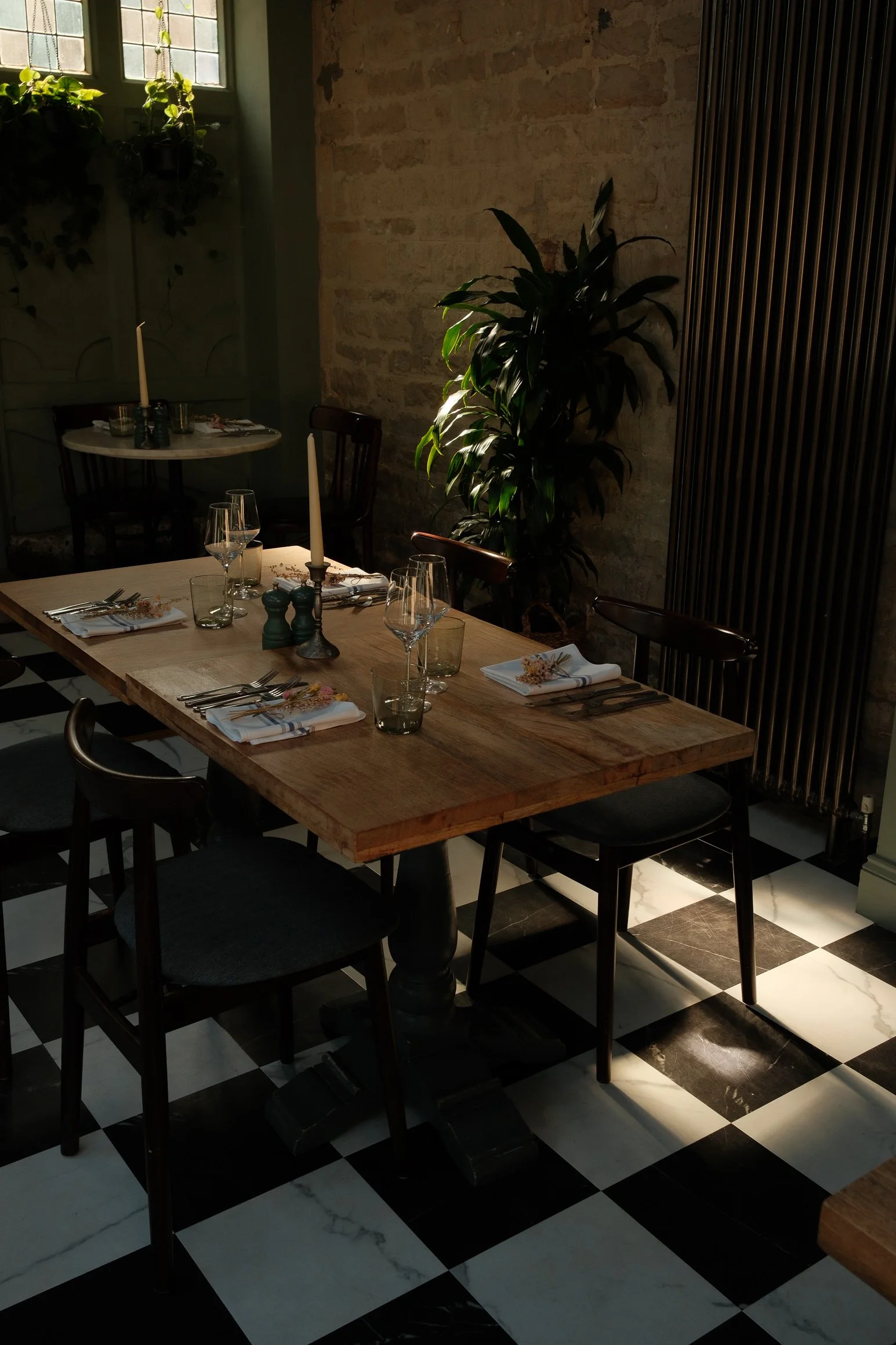 A cozy dining area with a wooden table set for a meal, featuring wine glasses, cutlery, napkins with floral decorations, and tall white candles in holders. There are black chairs around the table. The room has a black and white checkered floor, a bri