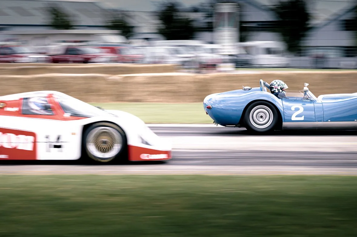 A white and red race car and a blue vintage race car moving at high speed on a race track