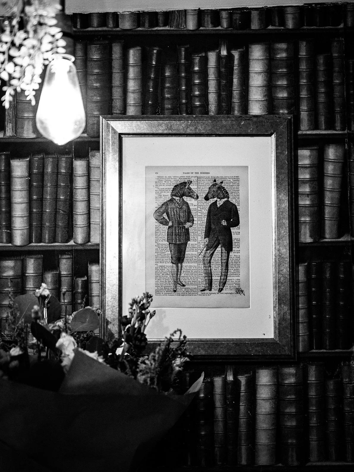 Black and white photo of two women with anthropomorphic bird heads walking and talking, framed and hanging on a wall of bookshelves, with flowers in the foreground.