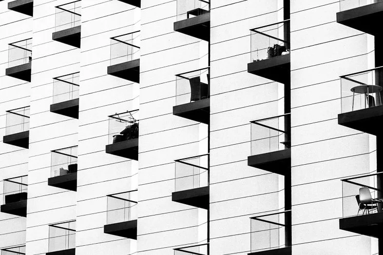Black and white photo of a modern apartment building with multiple balconies, each with a glass railing, some with furniture or plants visible.