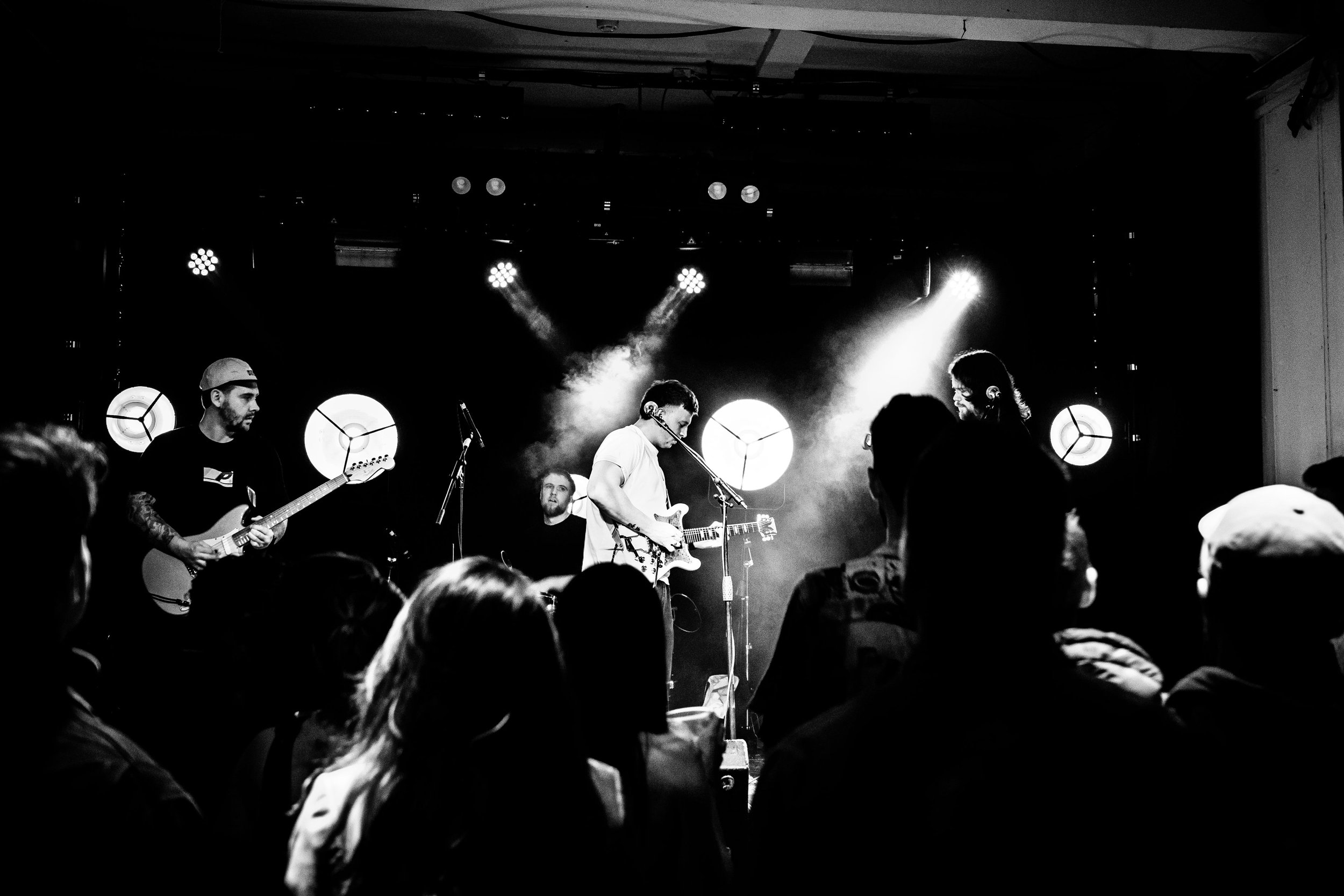 A live band performs on stage at a concert venue, with multiple musicians playing guitars and singing, while an audience watches.