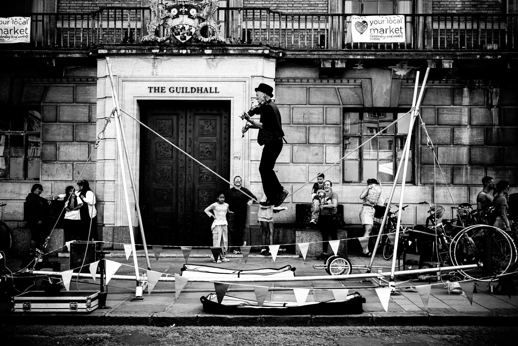 A street performer dressed as a clown standing on a slackline in front of a historic building labeled "The Guildhall". Several people, including children and adults, are watching and taking photos. Bicycles are parked nearby, and a crowd is gathered around the performance area with flags or bunting lines.