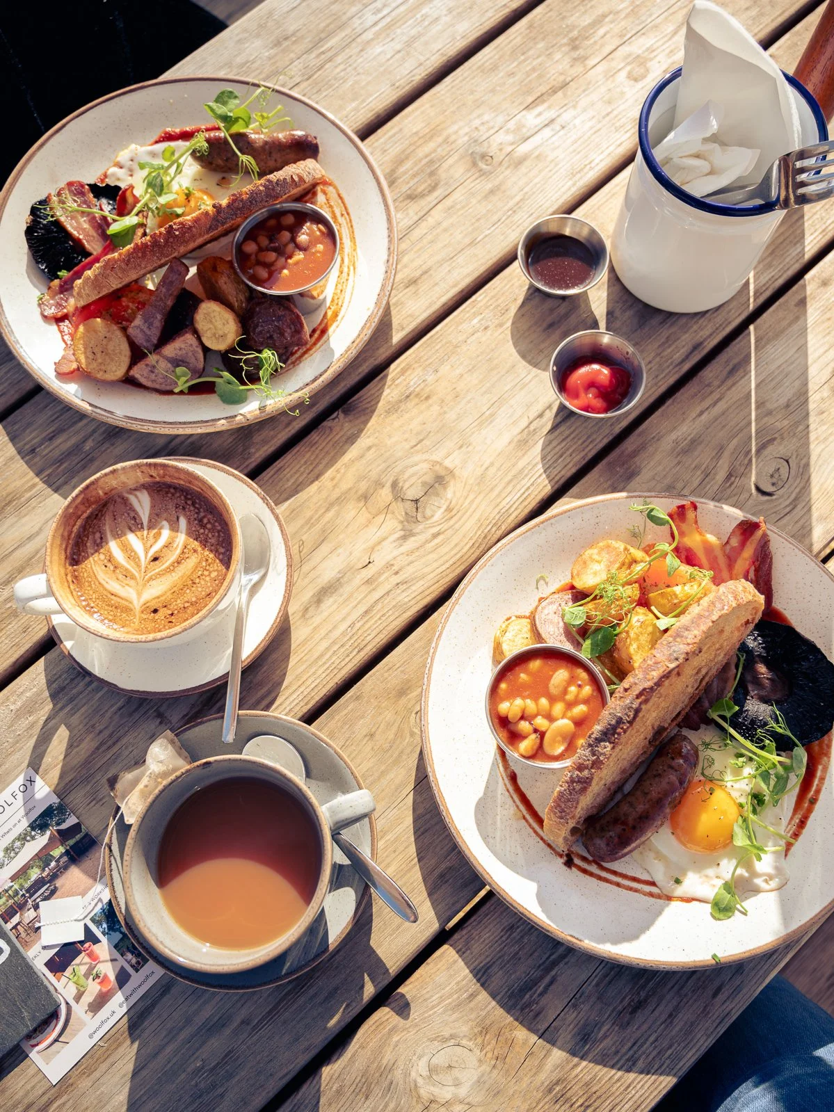 Two plates of breakfast food with fried eggs, sausages, beans, and toast, along with cups of coffee and tea on a wooden table outdoors.