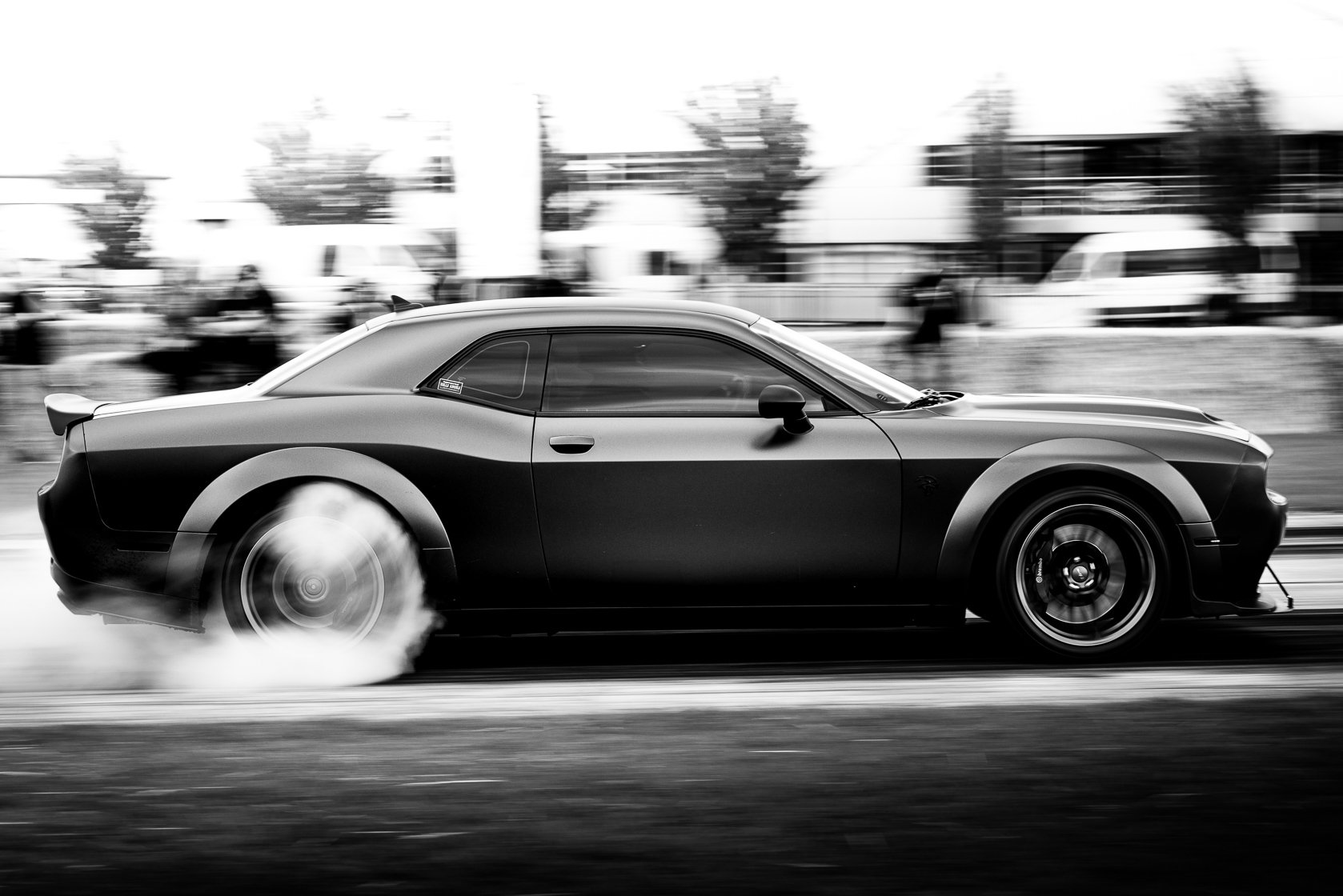 A black sports car performing a burnout, with smoke coming from the rear tires, in motion on a road in an urban area with blurred background of buildings and trees. The photo is in black and white.