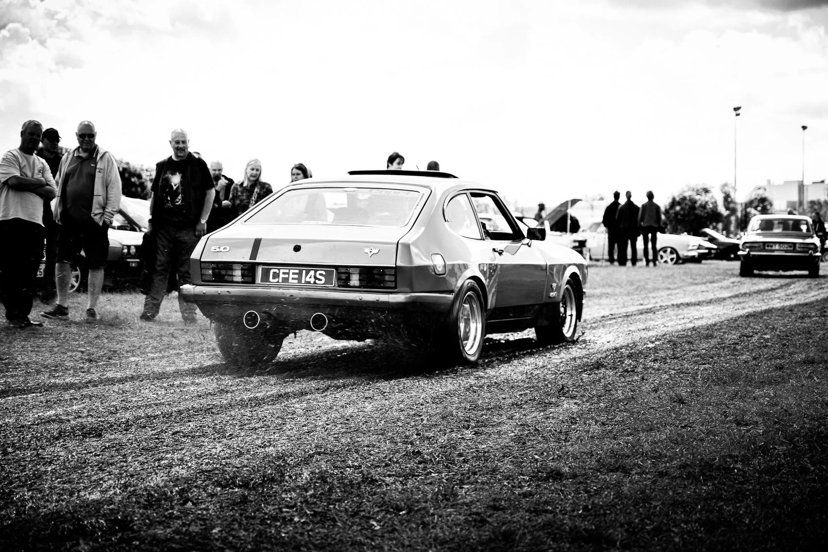A vintage racing car with a unique license plate CFE 14S on a gravel surface. Several people are standing nearby, with some of them looking at the car. Other classic cars are visible in the background on a cloudy day.