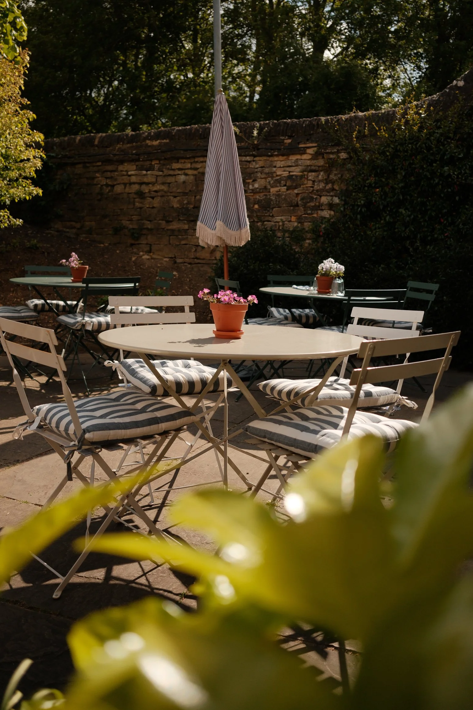 Empty outdoor patio with white and green tables and chairs, striped cushions, potted flowers, and a closed outdoor umbrella, surrounded by greenery and a brick wall.