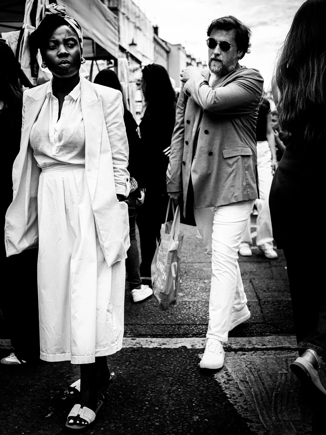 Black and white photo of a woman with a headwrap and white outfit walking in a busy street, alongside a man with sunglasses, a blazer, and white pants.