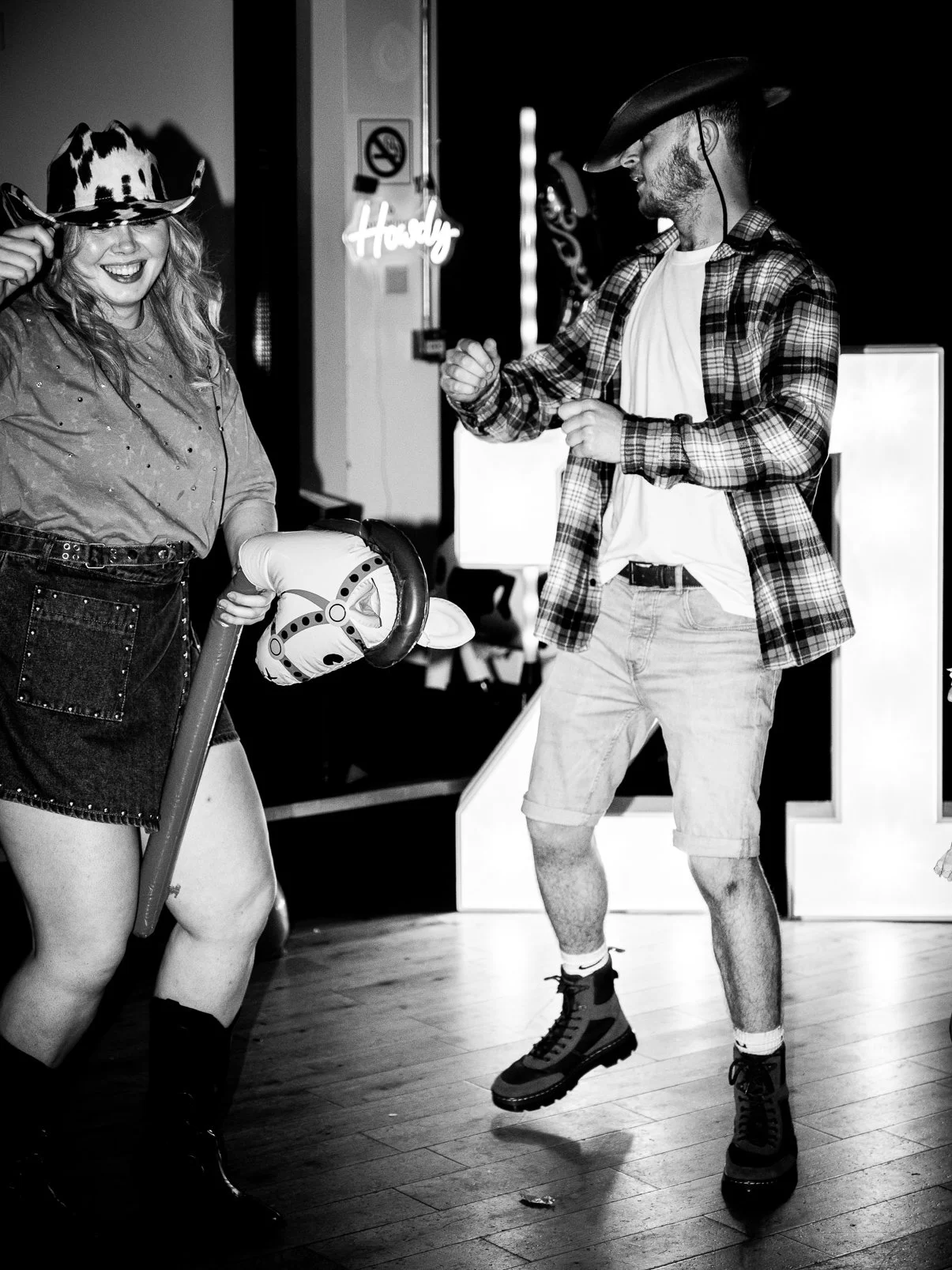 Two people dressed in cowboy-themed costumes dancing indoors, with one holding a toy horse and both wearing cowboy hats, in a black-and-white photo.