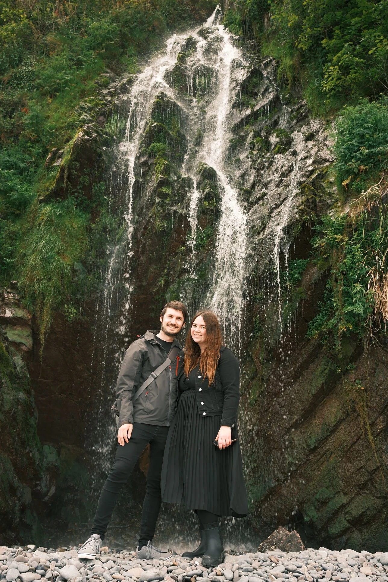 A couple stands on rocky terrain in front of a waterfall surrounded by greenery.