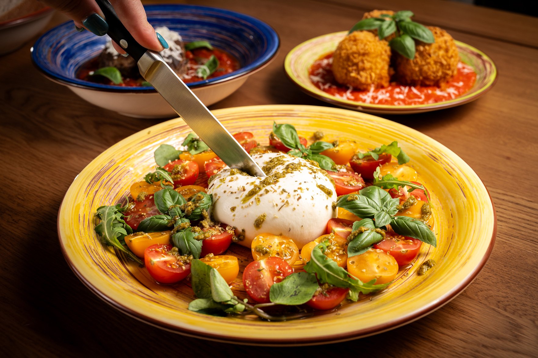 A colourful plate of cherry tomatoes, fresh basil, and a serving of burrata cheese garnished with herbs on a yellow plate, with two other dishes in the background on a wooden table.