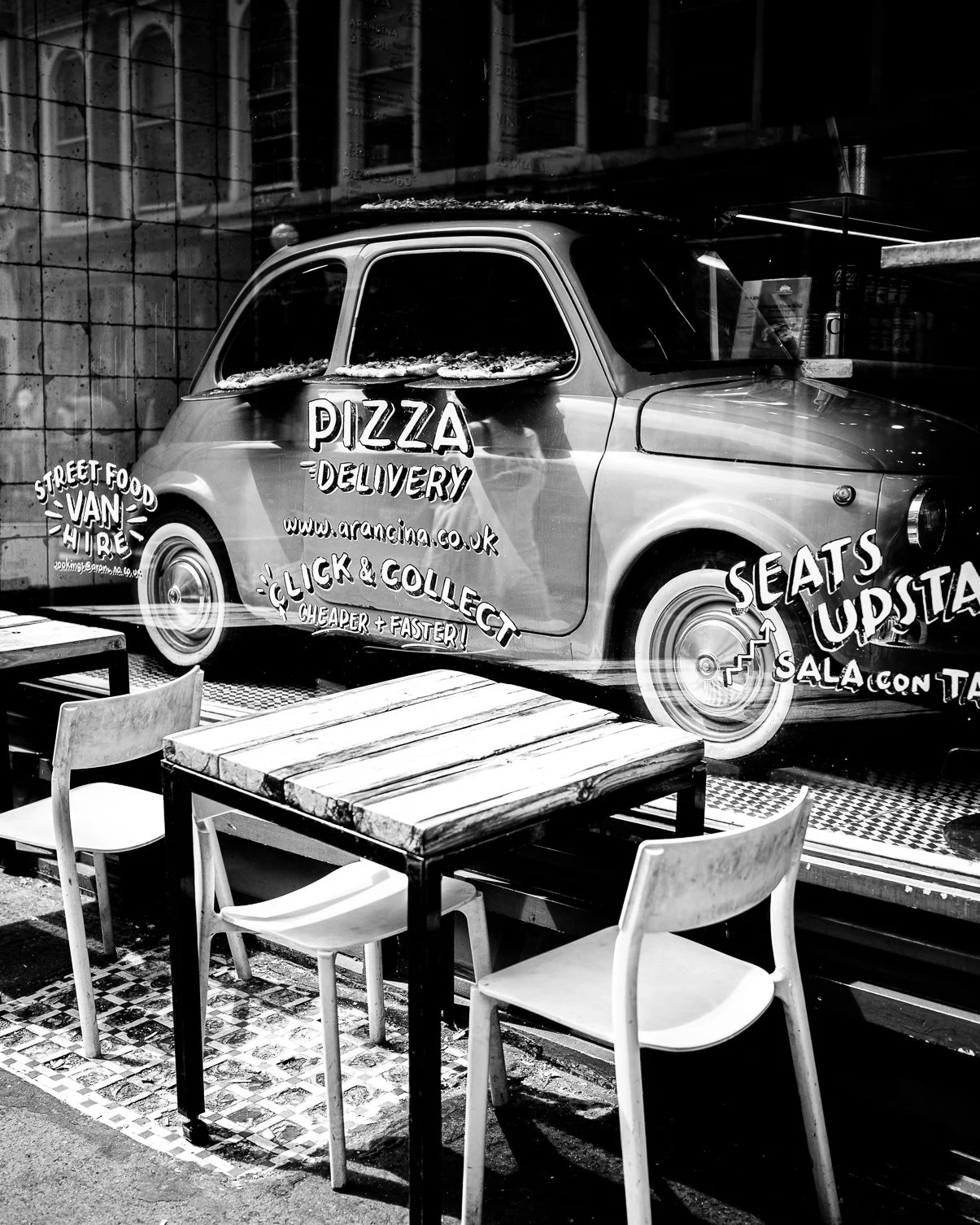 Black and white photo of a vintage car used as a pizza delivery van, with fun advertising text painted on the side, parked outside a restaurant with outdoor tables and chairs.