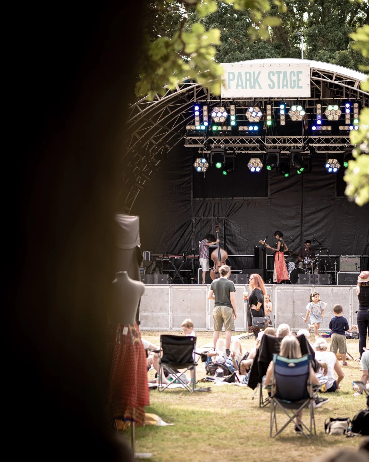 An outdoor concert stage labeled 'Park Stage' with musicians preparing to perform, and audience members sitting and standing on the grass watching and socializing.