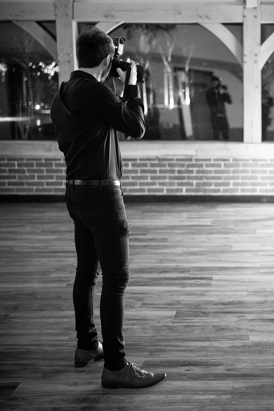 Photograph of a man taking a photo inside a room with wooden floors and brick walls, with his reflection visible in a large window behind him.