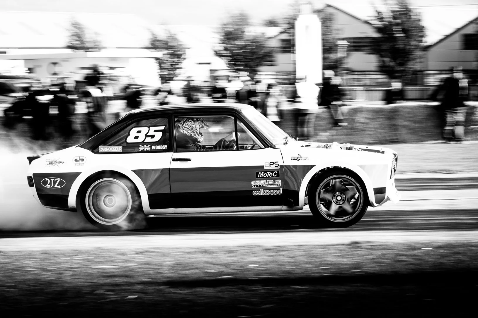 Black and white photo of a race car drifting during a race, with smoke coming from the rear tires. The car has the number 85 and various sponsor stickers on the side. The driver is wearing a helmet, and there are spectators blurred in the background.
